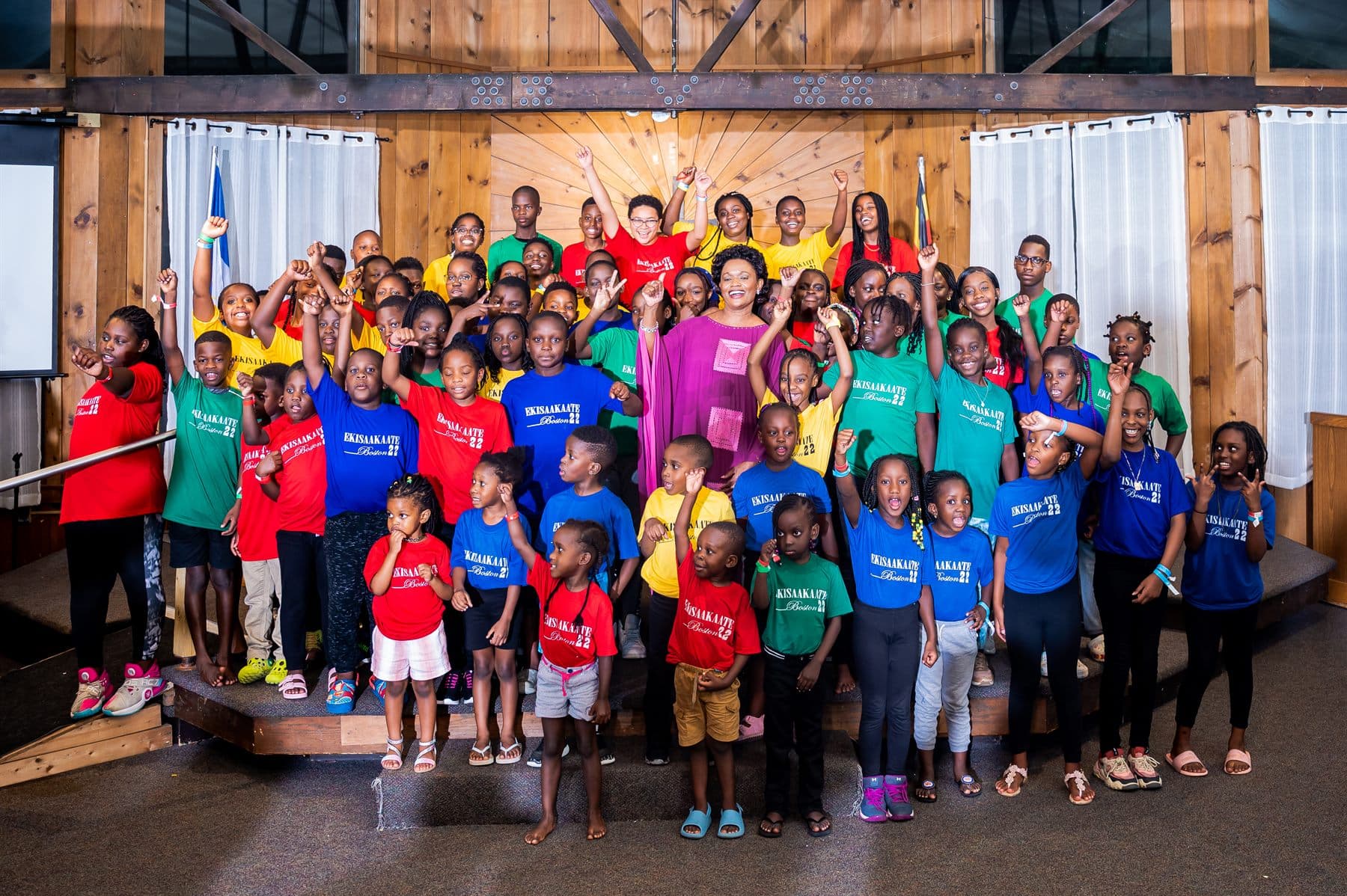 Ekisaakaate Boston campers in red, blue, green, and yellow team shirts gathered with Her Royal Highness the Nnaabagereka