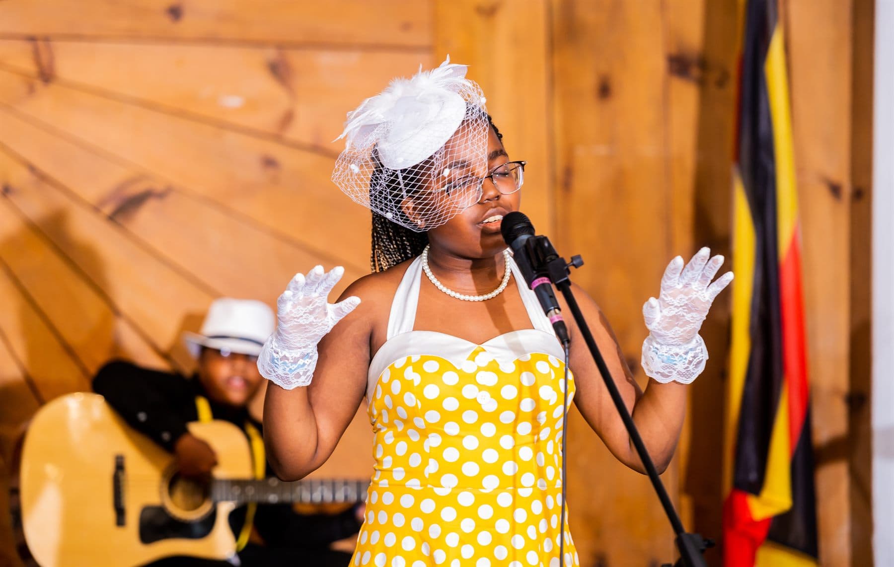 A young performer in a yellow polka-dot dress singing beside the Uganda flag